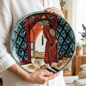 May include: A hand-painted ceramic plate with a Middle Eastern theme. The plate features a figure in a red robe, surrounded by turtles, within an arched doorway. The border is teal, and the background is black with blue and red accents.