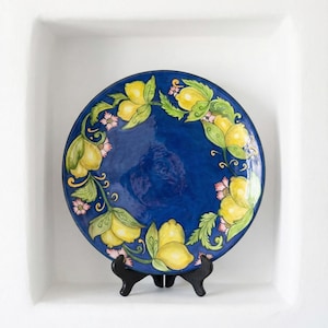 May include: A round, blue ceramic plate with a hand-painted lemon and floral design. The plate features yellow lemons, green leaves, and pink flowers arranged in a circular pattern. It is displayed on a black stand.