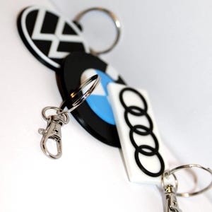 May include: Keychains featuring automotive logos, including BMW, Audi, and VW. The keychains have silver metal rings and clasps. The Audi keychain is white with black rings. The BMW keychain has a blue and white design.