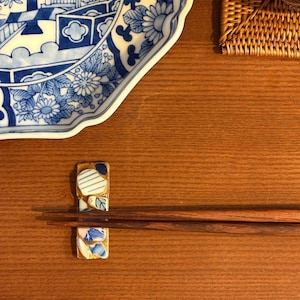 May include: A wooden table setting featuring a blue and white patterned plate, a woven placemat, and a decorative chopstick rest holding dark brown chopsticks. The chopstick rest has a mosaic design.