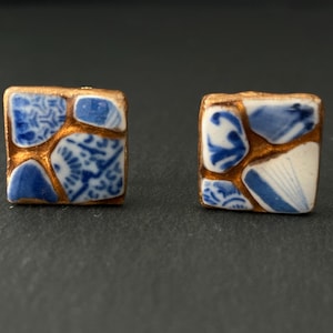May include: Square stud earrings with a mosaic design. Each earring features blue and white porcelain pieces set in a gold-colored metal frame. The porcelain has intricate patterns. The earrings are displayed on a dark surface.