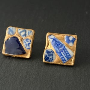 Könnte beinhalten: Ein Paar quadratische Ohrstecker mit goldfarbenem Rand. Jeder Ohrring zeigt ein Mosaik aus blau-weißen Keramikteilen. Ein Ohrring hat ein großes, dunkelblaues Stück, der andere ein federartiges Design.