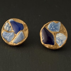Puede incluir: Un par de pendientes redondos con un borde dorado. Cada pendiente presenta un diseño de mosaico de piezas de cerámica azules y blancas. Las piezas de cerámica son de varios tonos de azul, algunas con patrones intrincados.
