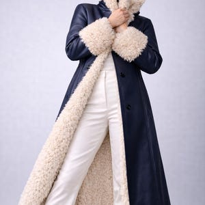 以下が含まれることがあります： クリーム色のフェイクファーの裏地とトリムが付いた、ネイビーブルーのロングコート。コートは、同じクリーム色のフェイクファーのハイカラーと袖口が特徴です。コートはボタンで留められ、白いパンツの上に着用されています。