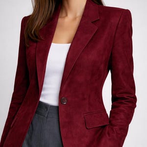 Puede incluir: Una blazer burdeos con cierre de un solo botón y solapa con muescas. La blazer tiene un corte entallado y un bolsillo en la parte delantera. La modelo lleva un top blanco y pantalones grises.
