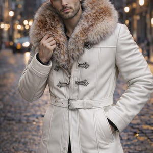 Könnte beinhalten: Ein cremefarbener Ledermantel mit Pelzkragen und dekorativen silbernen Verschlüssen. Der Mantel hat einen Gürtel in der Taille und Taschen. Das Model trägt eine schwarze Hose. Der Mantel ist ein stilvolles Oberbekleidungsstück.