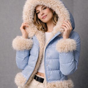 以下が含まれることがあります： ライトブルーのパファージャケット。フード、袖口、トリムにフェイクファーを使用。キルティングデザインでボタン留め。黒いベルトが見えます。