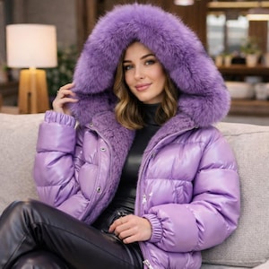 以下が含まれることがあります： 光沢のあるラベンダー色のパファージャケット。大きな紫色の毛皮トリムのフードが付いています。ジャケットはキルティングデザインで、ハイカラーです。モデルは黒いトップスと黒い革のパンツを着用しています。ジャケットはスタイリッシュなアウターウェアです。