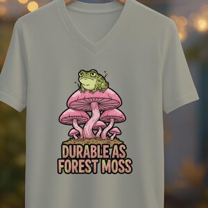 May include: A light gray V-neck t-shirt featuring a graphic of a green frog atop pink mushrooms. The text "DURABLE AS FOREST MOSS" is below the mushrooms. The shirt is hanging on a wooden hanger.