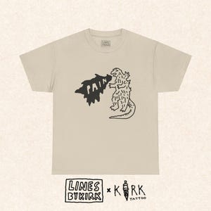 以下が含まれることがあります： ベージュ色のTシャツ。黒いグラフィックで、ゴジラのような生き物が「PAIN」という言葉を吐き出している。Tシャツはクルーネックで半袖。Tシャツの下部には「LINES BY KIRK」と「KIRK TATTOO」の文字が表示されている。