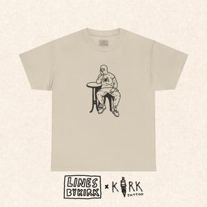 May include: Beige t-shirt featuring a black illustration of a person in a hooded outfit sitting at a small table. The person is resting their chin on their hand. The shirt has the text "LINES BY KIRK" and "KIRK TATTOO" at the bottom.