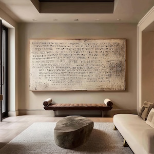 Puede incluir: Un salón con una gran obra de arte enmarcada con escritura antigua. Debajo, un banco de cuero marrón con cojines se encuentra contra una pared neutra. Una mesa de centro de piedra y un sofá crema completan la decoración de la habitación.