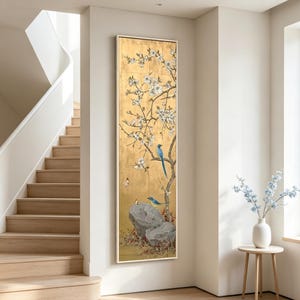 May include: A tall, rectangular wall art piece featuring a gold background with a painted tree branch bearing white blossoms. Two blue birds are perched on the branches and rocks, with small red flowers at the base and a grey stone in the foreground.