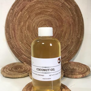 Puede incluir: Una botella transparente de aceite de coco con tapa y etiqueta blancas, colocada sobre un posavasos tejido. La etiqueta dice "Coconut Oil, Raw and Unrefined, Net Wt 500ml, Made in Nigeria". Un tapiz de pared tejido está en el fondo.