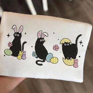 May include: White sweatshirt featuring embroidered black cats with bunny ears, Easter eggs, flowers, and small bunny figures. The design includes a cat emerging from a cracked egg. The design is in a cartoon style.