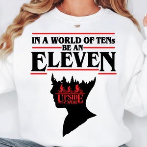 Peut inclure: Sweat-shirt blanc avec l'inscription "IN A WORLD OF TENS BE AN ELEVEN" en noir et rouge. Une silhouette noire d'une tête avec une forêt et des cyclistes est en dessous. Le mot "UPSIDE DOWN" est en rouge.
