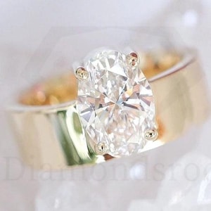 May include: A gold band engagement ring with a large, oval-cut diamond. The diamond is set in a four-prong setting, and the band is wide and polished. The ring is displayed on a white, textured surface.