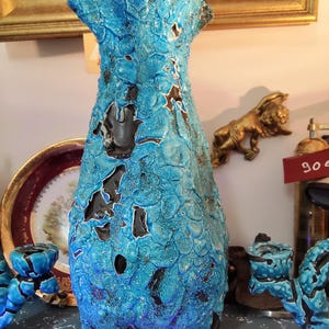 Könnte beinhalten: Eine hohe, strukturierte türkisfarbene Keramikvase mit einem einzigartigen, organischen Design. Die Vase hat eine raue, blasenartige Oberfläche mit dunkelbraunen Akzenten und Öffnungen. Es ist ein dekorativer Gegenstand, wahrscheinlich für die Inneneinrichtung.