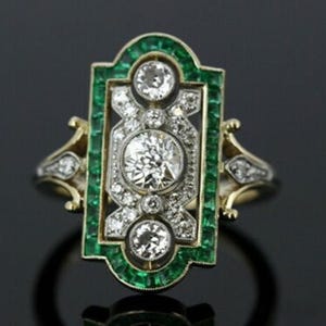 May include: An Art Deco style ring featuring a central, large, round diamond surrounded by smaller diamonds and emeralds. The ring has a rectangular emerald border and gold accents, creating a vintage aesthetic.