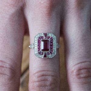 Puede incluir: Un anillo plateado con una gema rectangular de color rojo intenso en el centro, rodeada de piedras rojas y claras más pequeñas. El anillo tiene forma rectangular con un diseño detallado y ornamentado. El anillo está en un dedo.
