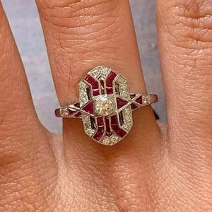 Puede incluir: Un anillo de estilo vintage con un diamante central y piedras preciosas rojas y blancas. El anillo tiene un diseño Art Déco con formas geométricas y una banda plateada. El anillo está en un dedo.