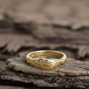 May include: A gold-colored ring featuring a design of two hands clasped together. The ring is set against a rustic, wooden background, highlighting its polished finish and intricate details. The ring's design symbolizes unity and connection.