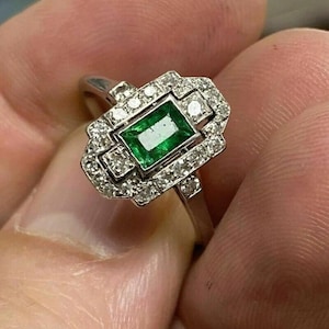 May include: A silver ring with a rectangular emerald centre stone, encircled by small diamonds. The ring has an Art Deco design with geometric details, showcasing a classic and elegant style.