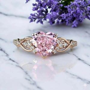 May include: A rose gold ring featuring a large, heart-shaped pink gemstone. The band has intricate, interwoven patterns and small, clear accent stones. The ring is set on a marble surface, with purple flowers in the background.