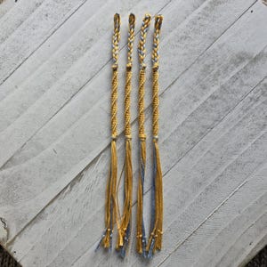 Puede incluir: Cinco pulseras de amistad hechas a mano con un diseño retorcido. Las pulseras son principalmente doradas con detalles azules y se exhiben sobre una superficie de madera blanca. Cada pulsera tiene un lazo trenzado en la parte superior y borlas en la parte inferior.