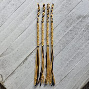 Puede incluir: Cinco pulseras de amistad hechas a mano con un diseño en espiral. Cada pulsera presenta hilos dorados y azul marino, con una parte superior trenzada y largas borlas colgantes. Las pulseras están dispuestas verticalmente sobre un fondo de madera blanca.