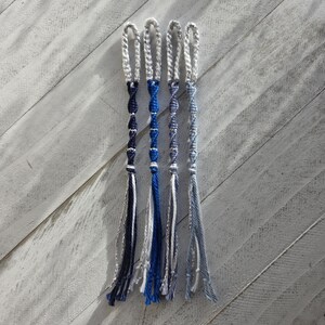 Puede incluir: Cuatro pulseras de amistad hechas a mano en tonos de azul, blanco y gris. Cada pulsera presenta una parte superior en bucle y un diseño trenzado o retorcido, con hilos colgantes en la parte inferior. Las pulseras están dispuestas sobre una superficie de madera gris claro.
