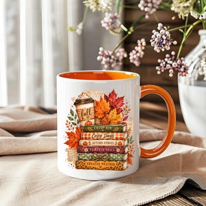 Könnte beinhalten: Ein weißer Keramikbecher mit orangefarbenem Innenraum und Henkel, verziert mit einer Grafik einer Kaffeetasse auf einem Stapel von fünf Büchern. Die Bücher tragen die Aufschriften Crisp Air, Cozy Days, Autumn Stories, Pumpkin Spice und Sweater Weather.