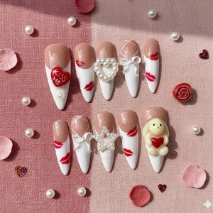 May include: A set of ten artificial nails with a variety of designs. The nails feature a white base with nude tips, adorned with red lips, bows, pearls, and a heart-shaped charm. One nail has a bunny holding a heart, and another says "Kiss Me".