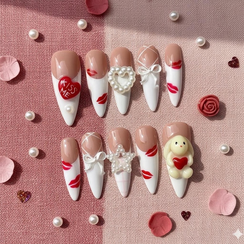 May include: A set of ten artificial nails with a variety of designs. The nails feature a white base with nude tips, adorned with red lips, bows, pearls, and a heart-shaped charm. One nail has a bunny holding a heart, and another says "Kiss Me".