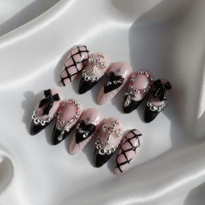May include: A set of almond-shaped press-on nails with a gothic theme. The nails feature a combination of black and pink colors, with intricate designs including crosses, bows, and lattice patterns. Some nails are adorned with rhinestones.