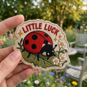 May include: An embroidered patch with a cream-colored background featuring a red ladybug with black spots on a green leaf. The text "LITTLE LUCK" is stitched in red above the ladybug, flanked by small yellow stars. White flowers with yellow centers are on the right side.