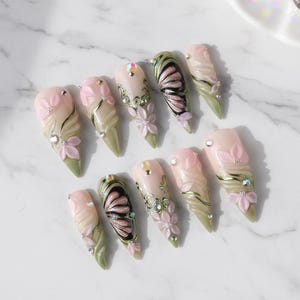 May include: A set of ten almond-shaped artificial nails. Each nail features a light pink base with intricate designs, including pink flowers, green accents, and sparkling rhinestones. Some nails showcase butterfly wing patterns.