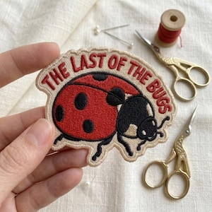 May include: An embroidered patch featuring a red ladybug with black spots on a cream background. The text "THE LAST OF THE BUGS" is stitched in red thread in an arc above the ladybug. The patch has a scalloped edge and is held by fingers.