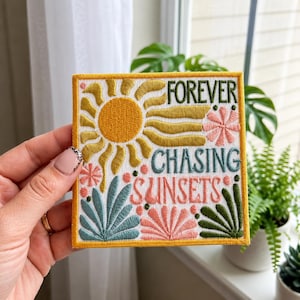 May include: A square embroidered patch featuring a yellow sun, pink and teal floral motifs, and green foliage. The text reads FOREVER CHASING SUNSETS in dark green and pink thread against a white background with a yellow border.