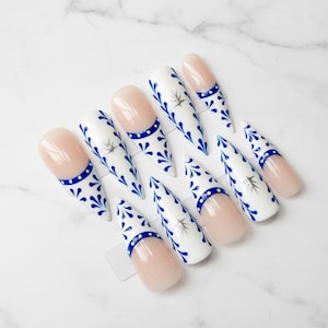 May include: A set of artificial nails with a white base, adorned with blue floral patterns and a blue band at the base. Some nails feature a nude-colored tip. The nails are arranged on a white marble surface.