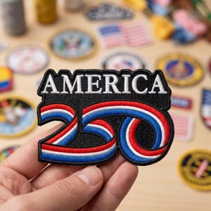 May include: An embroidered patch featuring the word AMERICA in white capital letters above the number 250. The numbers are stylized with a continuous ribbon design in red, white, and blue stripes against a solid black background.