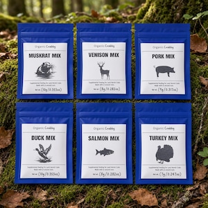 May include: Six blue pouches of Organic Crabby supplemental feeding for land hermit crabs. Each pouch features a white label with a black animal silhouette and text: Muskrat Mix 10g/0.353oz, Venison Mix 8g/0.282oz, Pork Mix 9g/0.317oz, Duck Mix 10g/0.353oz, Salmon Mix 8g/0.282oz, and Turkey Mix 7g/0.247oz.