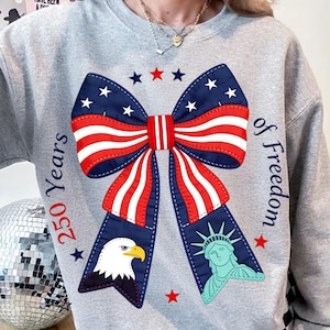 Op de afbeelding: Een grijze sweatshirt met een patriottisch strikdesign in rood, wit en blauw. Het ontwerp bevat een adelaar, het Vrijheidsbeeld en de tekst "250 Years of Freedom."