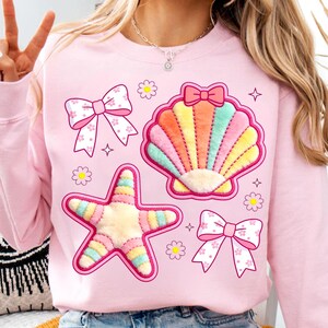 May include: Pink sweatshirt featuring embroidered seashell, starfish, and bow designs in pastel colors. The sweatshirt also has small flower and star details. A cute and whimsical design.