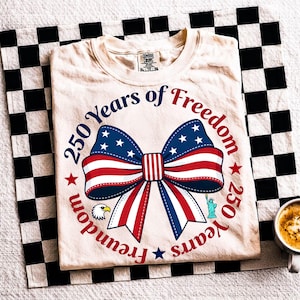 Puede incluir: Camiseta color crema con diseño patriótico. La camiseta presenta un gran lazo en rojo, blanco y azul, con estrellas y rayas, y el texto "250 Years of Freedom" en un patrón circular. También se ven un águila y la Estatua de la Libertad.