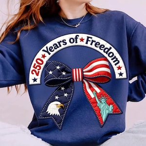 Könnte beinhalten: Marineblaues Sweatshirt mit patriotischem Schleifendesign. Die Schleife zeigt die amerikanische Flagge, einen Adler und die Freiheitsstatue. Der Text "250 Years of Freedom" ist bogenförmig angeordnet.