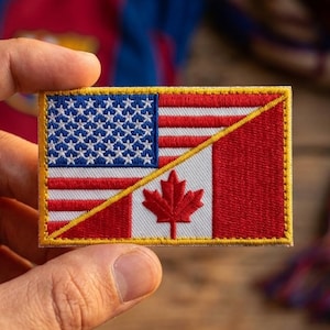 May include: A rectangular patch featuring the flags of the United States and Canada. The US flag is on the left, with the Canadian flag on the right. The patch has a gold border and is held in a person's hand.