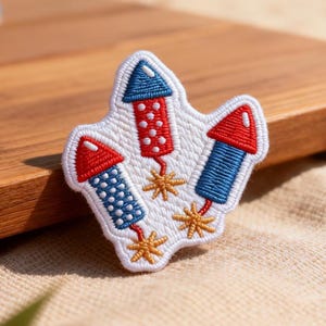 May include: Embroidered patch featuring three stylized fireworks in red, white, and blue. Each firework has a red or blue tip and is outlined in white. Gold starbursts are beneath the fireworks. The patch is on a tan surface.