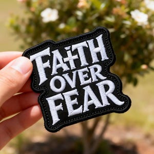 May include: A black patch with white embroidered text that reads "FAITH OVER FEAR." The patch has a black border and is held in a hand. The background is blurred, showing greenery and flowers.