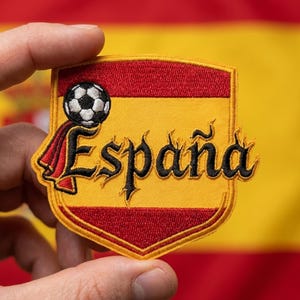 May include: Embroidered patch in the shape of a shield with the word "España" in black script. A soccer ball sits above the word, and the shield is red and yellow, with a red banner. The background is a Spanish flag.